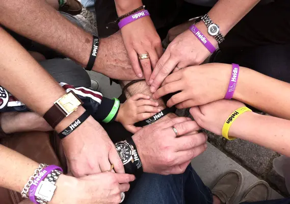 HeldenAusflug.de Hände aufeinander mit Armbändern mit Held/Heldin Aufschrift