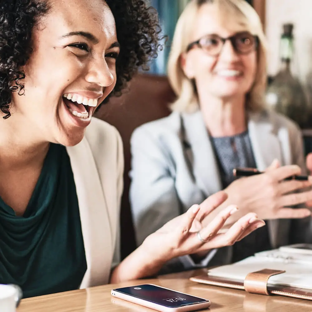 Zwei lachende Frauen sitzend am Tisch bei einem Meeting mit Notizheft und Handy.