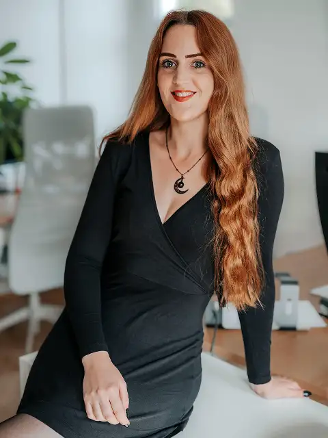 Portrait von Laura die sich an ein Regal lehnt und in die Kamera lacht.